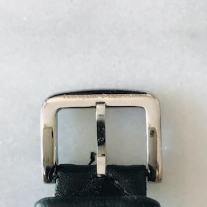May include: Close-up of a silver metal buckle with the engraved text "kate spade New York." The rectangular buckle has a central bar and is attached to a black leather strap.