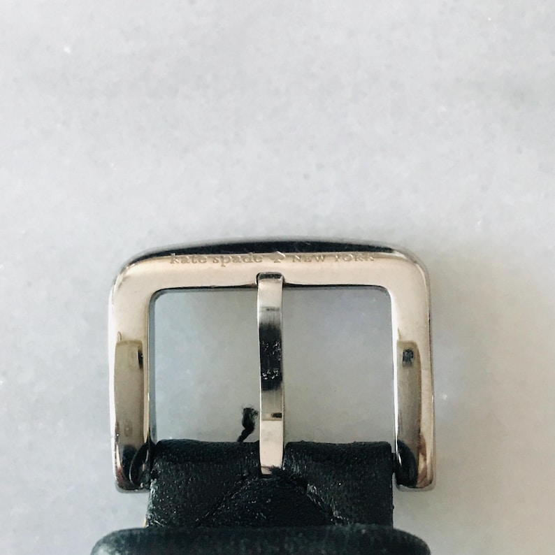 May include: Close-up of a silver metal buckle with the engraved text "kate spade New York." The rectangular buckle has a central bar and is attached to a black leather strap.