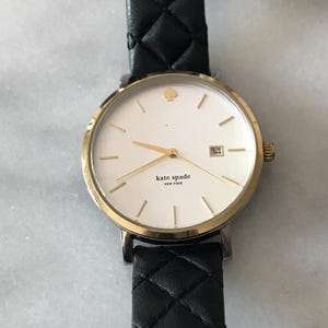 May include: A gold-toned Kate Spade watch with a white face and black leather strap. The watch features gold hands and markers, a date display, and the Kate Spade New York logo. The strap has a quilted pattern.