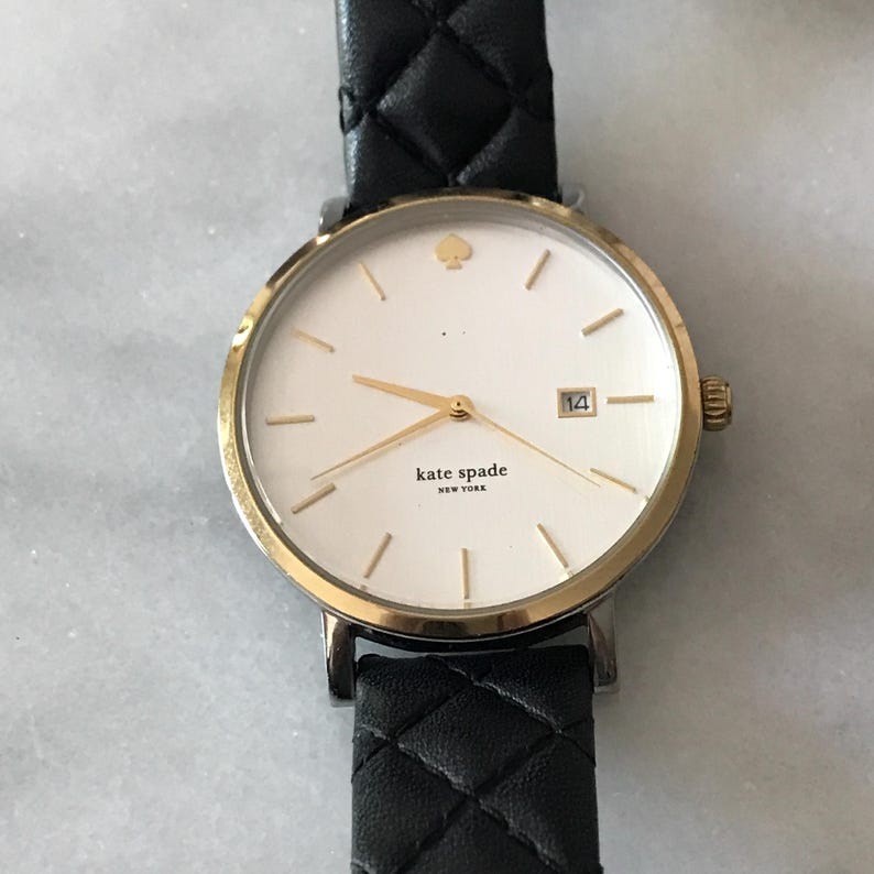 May include: A gold-toned Kate Spade watch with a white face and black leather strap. The watch features gold hands and markers, a date display, and the Kate Spade New York logo. The strap has a quilted pattern.