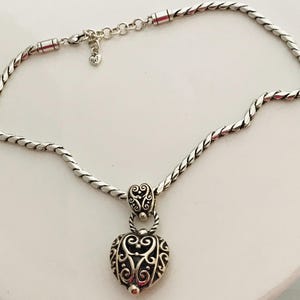 May include: A silver-toned necklace featuring a twisted rope chain and a heart-shaped pendant with intricate filigree scrollwork. The pendant is attached to the chain by a decorative bail, and the necklace includes a small heart-shaped charm.
