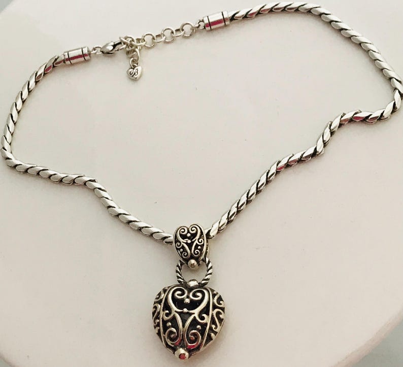 May include: A silver-toned necklace featuring a twisted rope chain and a heart-shaped pendant with intricate filigree scrollwork. The pendant is attached to the chain by a decorative bail, and the necklace includes a small heart-shaped charm.
