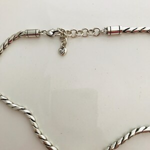 May include: A silver-toned necklace featuring a twisted rope-style chain. The piece includes a lobster claw clasp, a series of circular adjustment links, and a small heart-shaped charm dangling from the clasp, which is embossed with the letter B.