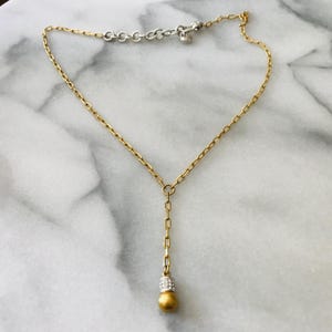 Puede incluir: Un collar en forma de Y de tono dorado con una cadena fina de eslabones rectangulares. El colgante central termina en una cuenta esf&eacute;rica dorada, rematada por una peque&ntilde;a banda plateada incrustada con diminutos cristales transparentes.