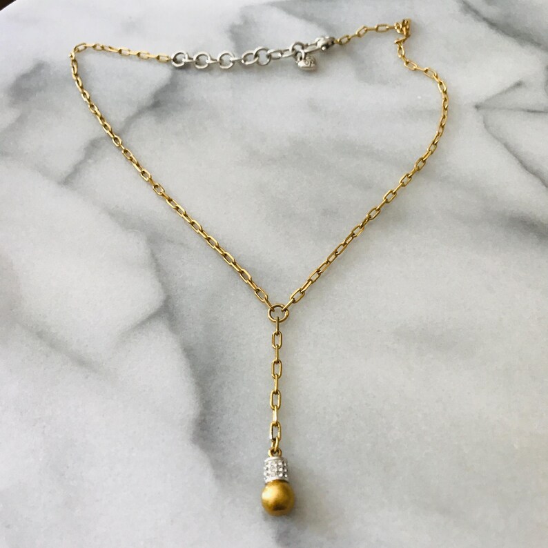 Puede incluir: Un collar en forma de Y de tono dorado con una cadena fina de eslabones rectangulares. El colgante central termina en una cuenta esf&eacute;rica dorada, rematada por una peque&ntilde;a banda plateada incrustada con diminutos cristales transparentes.
