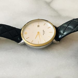 May include: A silver and gold-tone Kate Spade watch with a white face, gold hands, and a black leather strap. The watch face has a small gold spade logo and the words "kate spade new york". The strap is quilted.