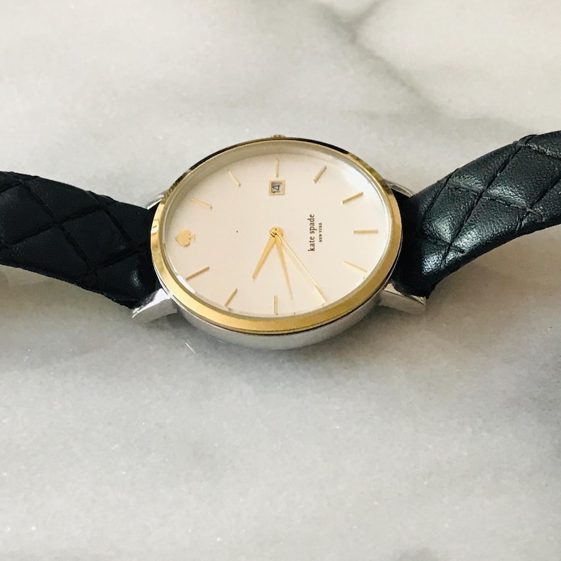 May include: A silver and gold-tone Kate Spade watch with a white face, gold hands, and a black leather strap. The watch face has a small gold spade logo and the words "kate spade new york". The strap is quilted.