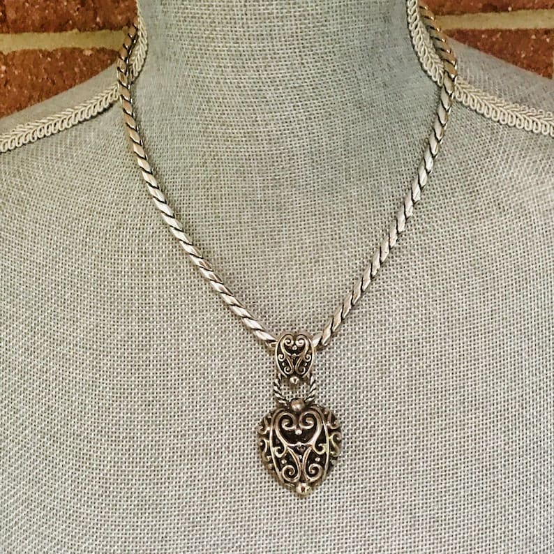 May include: A silver-toned necklace featuring a thick, twisted rope-style chain and a heart-shaped pendant. The pendant displays an intricate, openwork filigree design with swirling patterns, suspended from a small, matching decorative bail.