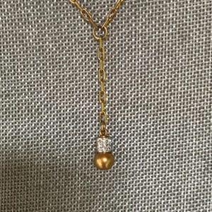 Puede incluir: Un collar de tono dorado con una cadena vertical que termina en un peque&ntilde;o colgante esf&eacute;rico de tono dorado. El colgante est&aacute; acentuado con una banda de tono plateado incrustada con peque&ntilde;as piedras claras y reflectantes en la parte superior.