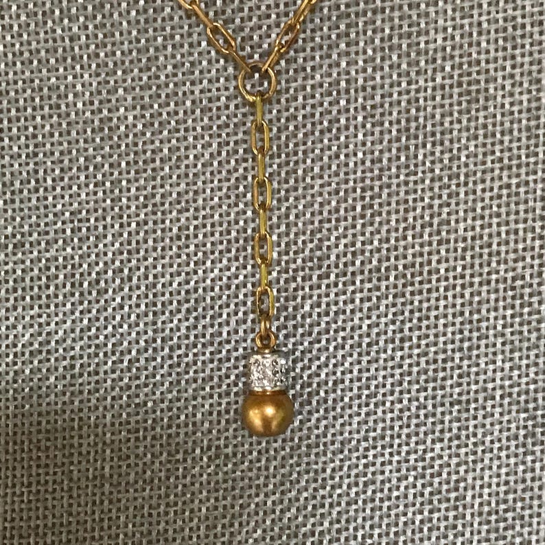 Puede incluir: Un collar de tono dorado con una cadena vertical que termina en un peque&ntilde;o colgante esf&eacute;rico de tono dorado. El colgante est&aacute; acentuado con una banda de tono plateado incrustada con peque&ntilde;as piedras claras y reflectantes en la parte superior.
