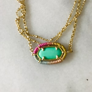May include: Gold-toned necklace with a layered chain design. The focal point is an oval pendant with a light green stone, surrounded by a colorful woven border of pink, yellow, and blue threads. The necklace is displayed on a white surface.