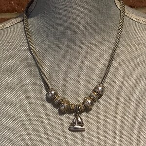 May include: A silver-toned necklace featuring a woven chain and a series of metallic beads in alternating silver and gold tones. A small, polished silver-toned sailboat charm hangs from the centre of the bead arrangement, creating a nautical-themed piece of jewellery.