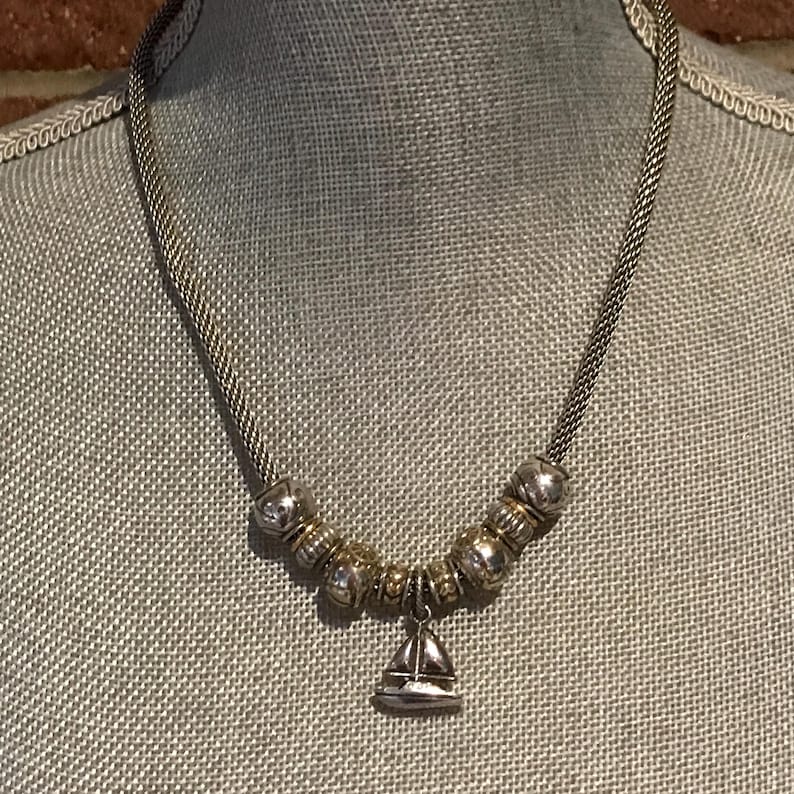 May include: A silver-toned necklace featuring a woven chain and a series of metallic beads in alternating silver and gold tones. A small, polished silver-toned sailboat charm hangs from the centre of the bead arrangement, creating a nautical-themed piece of jewellery.