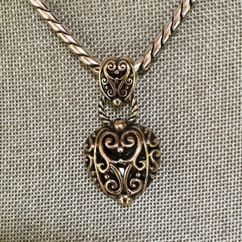 May include: A silver-toned necklace featuring a heart-shaped pendant with intricate scrollwork and filigree detailing. The pendant is suspended from a twisted rope-style chain, showcasing a polished metallic finish with darkened recessed areas.