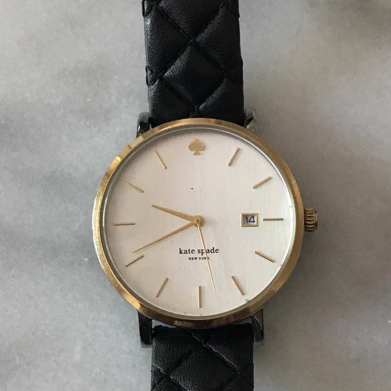 May include: A gold-tone Kate Spade watch with a white face, featuring a small spade logo and the text "kate spade NEW YORK". The watch has gold hands and markers, a date window at the 3 o'clock position, and a black quilted leather strap.