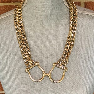 May include: A gold-toned necklace featuring two thick, interlocking curb chain strands that connect at the center to two polished, U-shaped metal loops. The chain links are uniform in size and have a reflective, metallic finish throughout the piece.