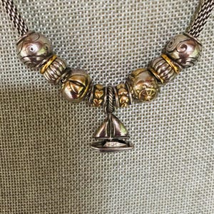 May include: A silver-toned chain necklace featuring a series of decorative beads in silver and gold tones, including textured spheres and heart-shaped spacers. A central sailboat-shaped charm hangs from the chain, detailed with small clear stones.
