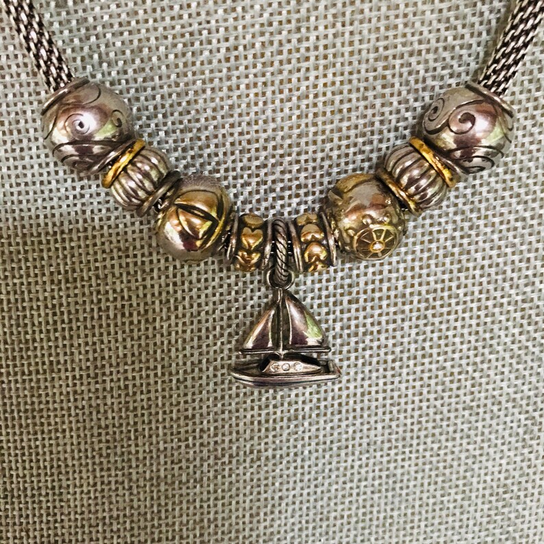 May include: A silver-toned chain necklace featuring a series of decorative beads in silver and gold tones, including textured spheres and heart-shaped spacers. A central sailboat-shaped charm hangs from the chain, detailed with small clear stones.