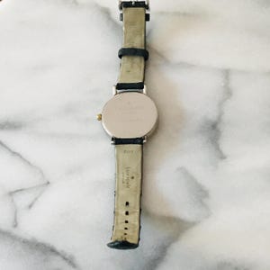 May include: A silver-toned Kate Spade watch with a black leather strap. The watch has a round face and a gold-coloured crown. The strap is marked "China" and "Kate Spade".
