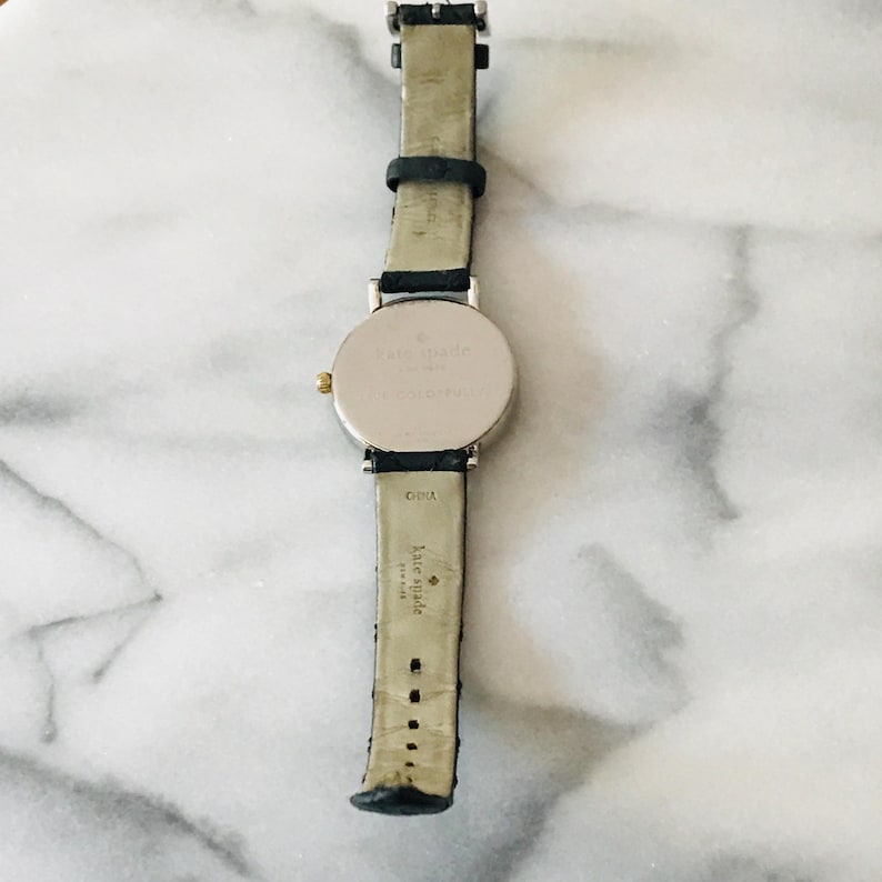 May include: A silver-toned Kate Spade watch with a black leather strap. The watch has a round face and a gold-coloured crown. The strap is marked "China" and "Kate Spade".