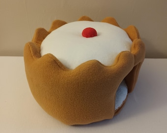 May include: A brown and white plush pet bed shaped like a cake with a red cherry on top. The bed has a small opening on the side for pets to enter.