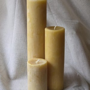 May include: Three natural beeswax pillar candles of varying heights. The candles are a warm yellow color and have a slightly textured surface. Each candle has a white wick at the top. The candles are arranged on a white fabric background.