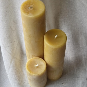 May include: Three natural beeswax pillar candles of varying heights. The candles are a warm yellow color and have a smooth, cylindrical shape. Each candle has a white wick centered on the top. The candles are arranged on a neutral-colored fabric.