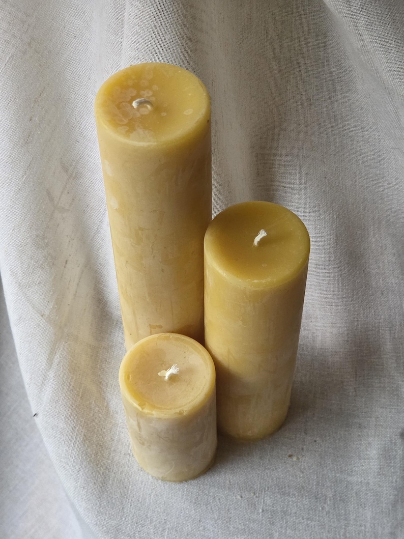 May include: Three natural beeswax pillar candles of varying heights. The candles are a warm yellow color and have a smooth, cylindrical shape. Each candle has a white wick centered on the top. The candles are arranged on a neutral-colored fabric.