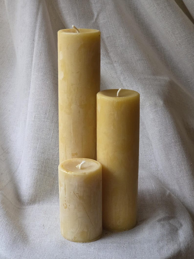 May include: Three natural beeswax pillar candles of varying heights. The candles are a warm yellow color and have a slightly textured surface. Each candle has a white wick at the top. The candles are arranged on a white fabric background.