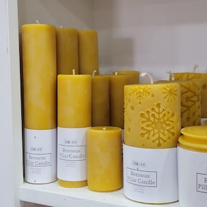 May include: A collection of yellow beeswax pillar candles. Some candles are cylindrical, while others have snowflake designs carved into the wax. Each candle sits on a white label that reads "Farm Hive Beeswax Pillar Candle".