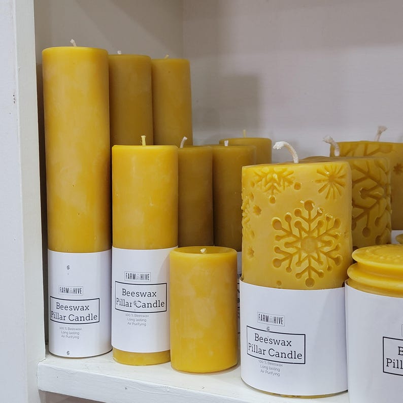 May include: A collection of yellow beeswax pillar candles. Some candles are cylindrical, while others have snowflake designs carved into the wax. Each candle sits on a white label that reads "Farm Hive Beeswax Pillar Candle".