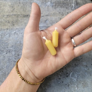 May include: Two small, cylindrical yellow candles with white wicks are held in a hand. The candles are made of a solid, waxy material. A gold bracelet with square beads and a silver ring are also visible.