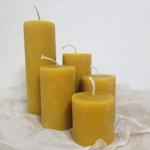 May include: Five yellow pillar candles of varying heights. The candles have white wicks and a smooth, solid appearance. The candles are arranged on a white cloth.