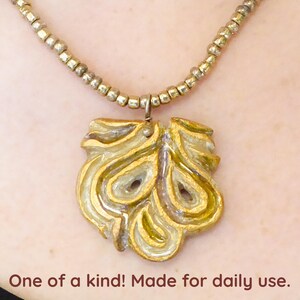 May include: A gold necklace with a unique, abstract pendant made of ceramic. The pendant has a swirling, organic design with a textured surface.