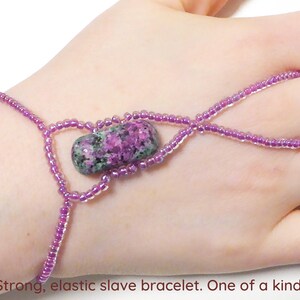 Op de afbeelding: Een paarse elastische slavenarmband met een roze en groene edelsteen. De armband is gemaakt van kleine kralen en heeft een uniek ontwerp.