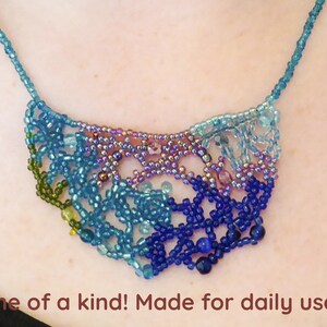 Puede incluir: Un collar de cuentas de colores con un diseño degradado en tonos de verde, azul y morado. El collar está hecho de pequeñas cuentas de semillas y tiene un patrón delicado e intrincado.