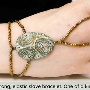 Könnte beinhalten: Ein elastisches Sklavenarmband in Gold und Grün mit einem großen, weißen, grünen und goldenen Anhänger mit Wirbelmuster.