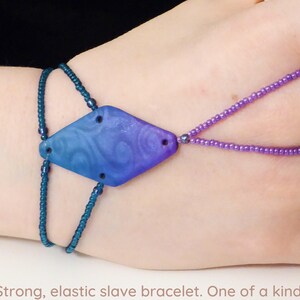 May include: A blue and purple slave bracelet with a diamond-shaped centerpiece. The centerpiece has a swirling pattern and is connected to the wrist and finger by beaded strands. The bracelet is a unique, one-of-a-kind piece.