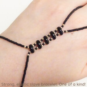 May include: Black beaded slave bracelet with gold accents. The bracelet is elastic and has a finger loop.