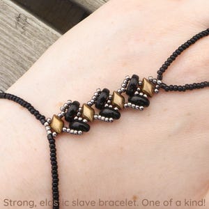 May include: Black and gold beaded bracelet with an elastic band. The bracelet is designed with a geometric pattern and is worn on a person's wrist.