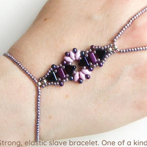 May include: A purple and pink beaded slave bracelet with a flower design. The bracelet is elastic and has a finger loop.