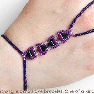 May include: A beaded slave bracelet with a dark purple and black design. The bracelet features a central section with black and purple geometric beads, connected by a beaded chain to a finger loop. The text at the bottom says "Strong, elastic slave bracelet. One of a kind!"