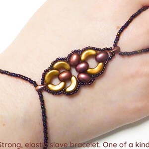 May include: A gold and purple beaded bracelet with an intricate design. The bracelet is elastic and has a finger loop.