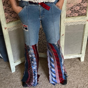 Pode incluir: Jeans azul de sino de ganga com um padr&atilde;o de gravata vermelha e branca na parte inferior das pernas. Os jeans t&ecirc;m um cinto vermelho com as palavras "Only the Strong" impressas nele.