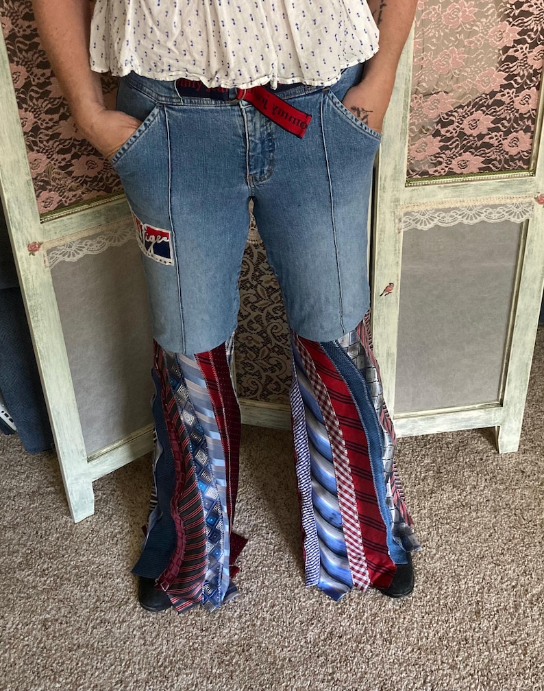 Pode incluir: Jeans azul de sino de ganga com um padr&atilde;o de gravata vermelha e branca na parte inferior das pernas. Os jeans t&ecirc;m um cinto vermelho com as palavras "Only the Strong" impressas nele.