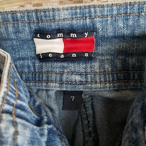 Pode incluir: Um par de cal&ccedil;as de ganga azuis com uma etiqueta Tommy Jeans e uma etiqueta de tamanho 7.