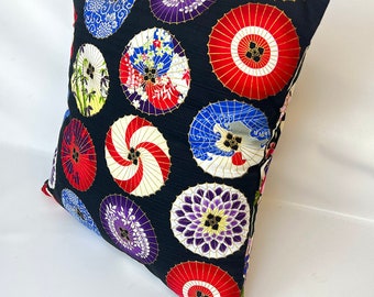 May include: Square decorative pillow with a black background and a pattern of colorful Japanese parasols. The parasols are red, blue, purple, and white with gold accents. The pillow is angled, showing its shape and design.