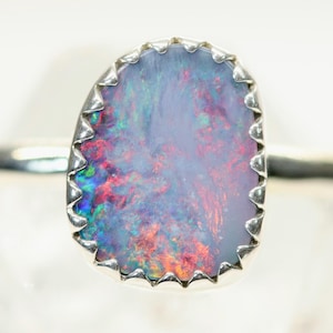 May include: A silver ring featuring an oval-shaped opal gemstone. The opal displays a mix of colors, including red, blue, and green, set within a scalloped bezel. The band is thin and polished.
