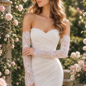 Op de afbeelding: Een strapless ivoorkleurige trouwjurk met een hartvormige halslijn, diagonaal geplooid lijfje en een rok tot op de grond. De jurk wordt gecombineerd met afneembare lange witte kanten handschoenen met ingewikkelde bloemmotieven en geschulpte randen.