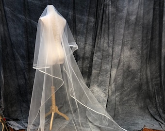 May include: A long, sheer white wedding veil featuring a delicate satin ribbon trim along the entire curved edge. The veil is draped over a mannequin, showcasing its flowing, floor-length design and lightweight, translucent tulle fabric construction.